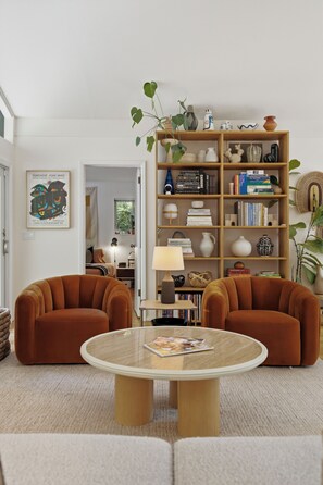 Ein Wohnzimmer mit zwei orangefarbenen Sessel, einem runden Couchtisch und einem Bücherregal mit Büchern und Dekoration.