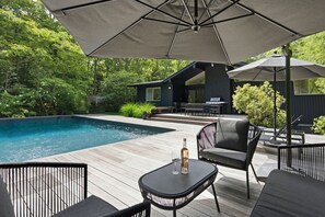 Eine Holzterrasse mit Gartenmöbeln, einem Pool und einem Haus im Hintergrund.