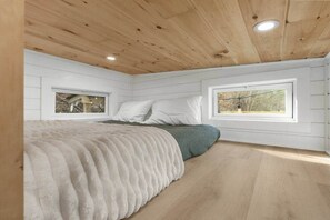 Ein Schlafzimmer mit Holzbalkendecke, zwei Fenstern, einem Bett mit weißer und grüner Decke sowie Holzboden.