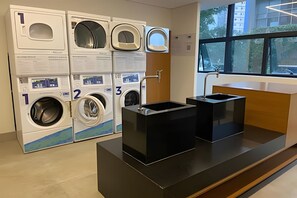 A modern laundry facility with multiple washing machines, a sink, and a window with a view of buildings. - Studio novíssimo próximo à Paulista (São Paulo)
