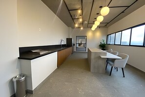Une cuisine moderne avec un plan de travail noir, un évier et un réfrigérateur. On y trouve également un coin repas avec une table et des chaises blanches. La pièce est dotée de grandes fenêtres et offre une vue sur le ciel.