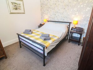 A neatly made bed with a checkered bedspread, two bedside lamps, a nightstand, and a floral wallpaper. - Halwyn Farm (Helston)