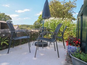 An outdoor seating area with metal chairs and a table, surrounded by greenery and a stone wall. - Halwyn Farm (Helston)