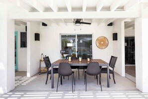 An outdoor dining area with a wooden table and chairs, a ceiling fan, and a large window with a view. - Adorable 4-bedroom house in La Quinta with private pool and mountain views! (La Quinta)