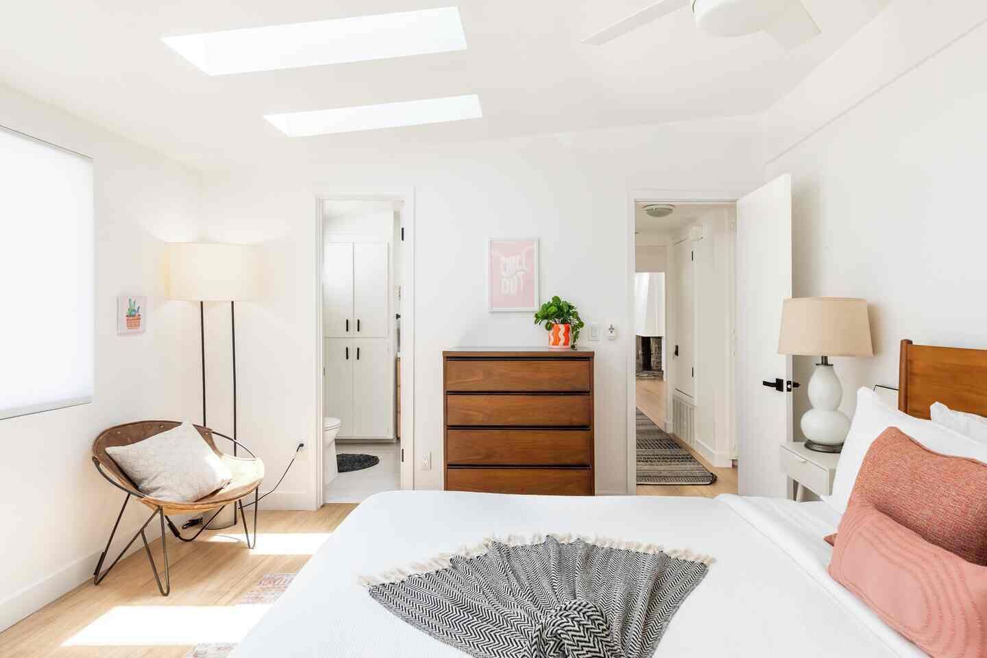 A bedroom with a bed, bedside table, lamp, dresser, chair, and a view of a hallway leading to a bathroom.
