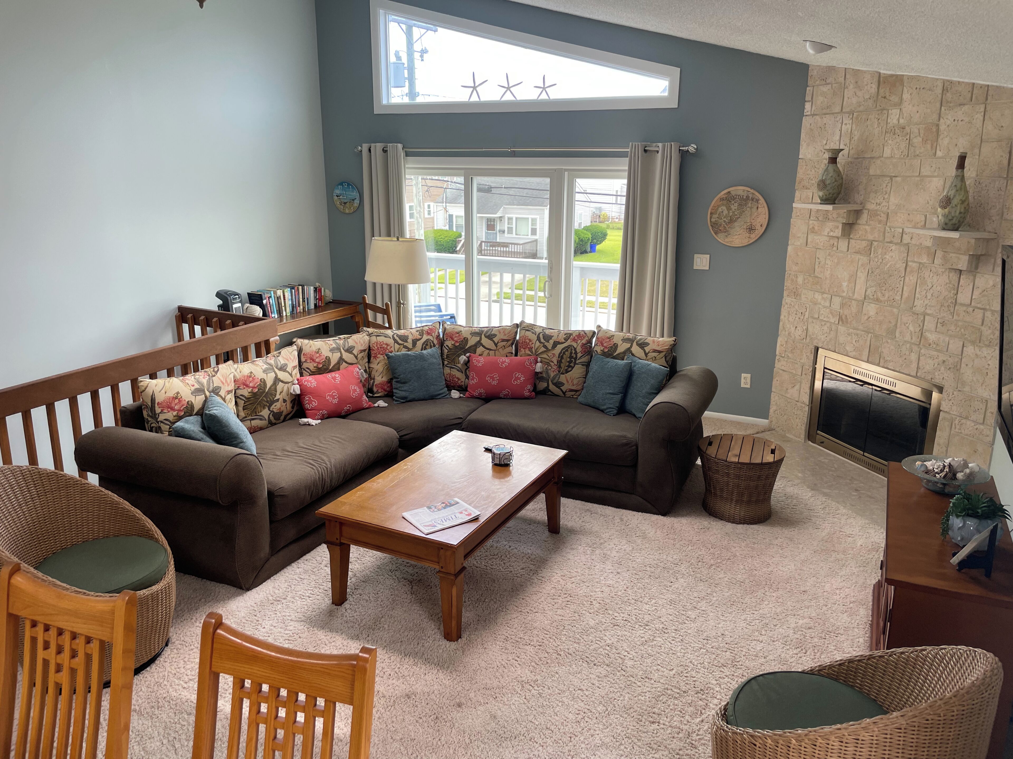 A living room with a sectional sofa, a coffee table, wicker chairs, a fireplace, and a view through a large window.