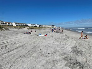 Ein Sandstrand mit Liegestühlen und Sonnenschirmen, einige Personen sowie Gebäude im Hintergrund.