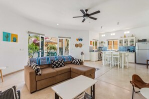 Ein modernes Wohnzimmer mit einer Couch, einem Esstisch und einer Küchenzeile mit weißen Schränken und einem Kühlschrank.