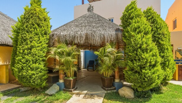 A courtyard with a thatched-roof structure, potted plants, and a pathway leading to a building entrance. - Cerritos Beach Poolside Retreat / 2BR + BBQ (El Pescadero)