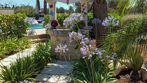 Un sendero en un jardín que lleva a una zona de piscina con un arco de piedra y plantas con flores.