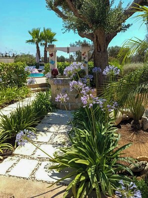 Ein Gartenpfad führt zu einem Poolbereich mit einem steinernen Bogen und blühenden Pflanzen.