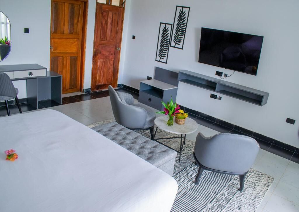 A modern hotel room with a bed, two grey armchairs, a small round table with flowers, a TV mounted on the wall, and a wooden door.