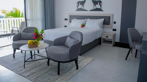 Una habitació d'hotel moderna amb un llit, dues butaques, una taula petita amb un ram de flors, una tauleta de nit i un balcó amb vistes.