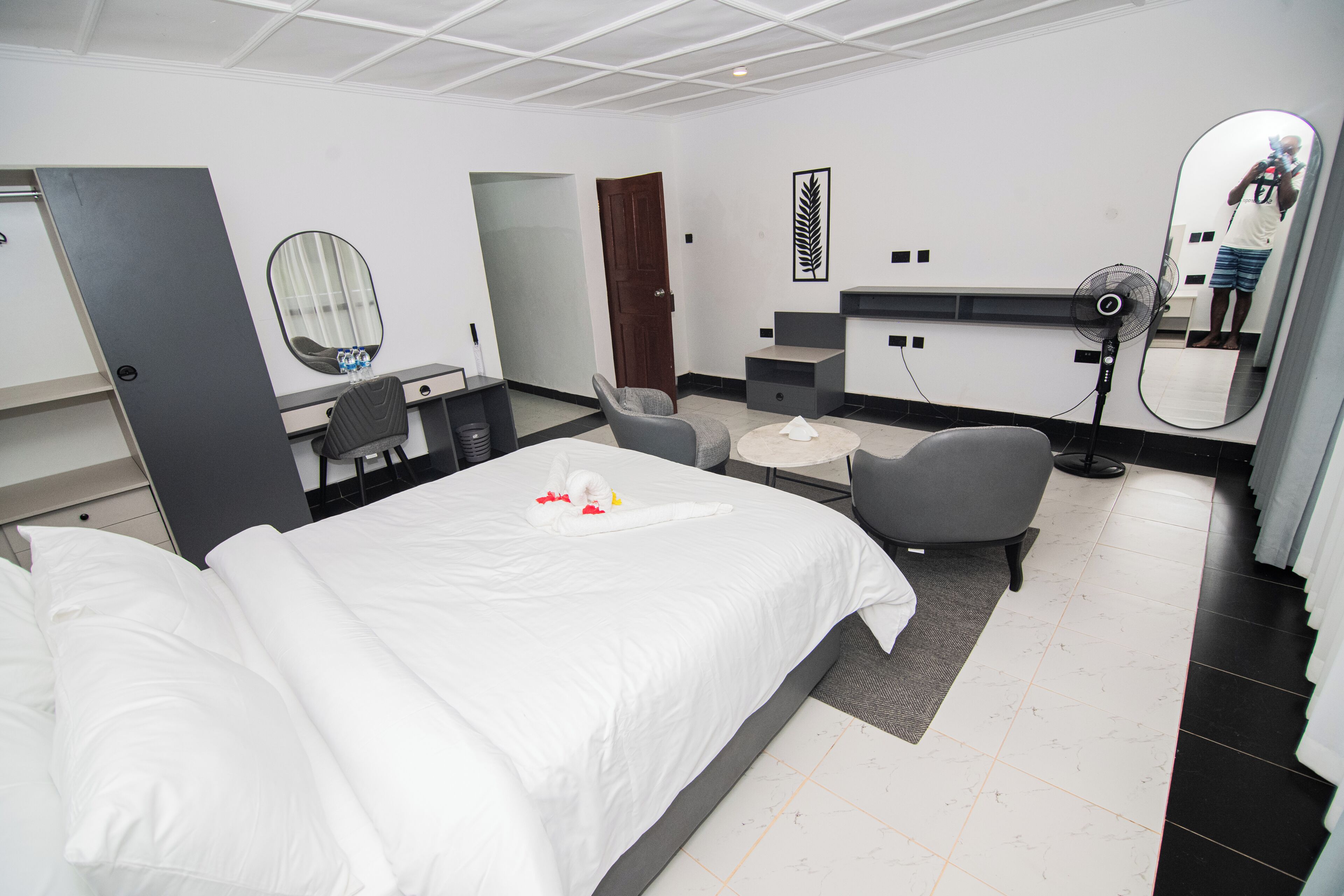 A hotel room with a bed, a desk, a chair, a mirror, and a fan.