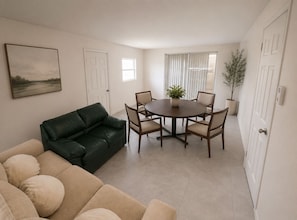 Ein Wohnzimmer mit einem grünen Ledersofa, einem beigefarbenen Sofa und einem runden Esstisch mit Stühlen. Auf dem Tisch steht ein Blumentopf und an der Wand hängt ein Gemälde.