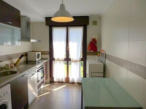A compact kitchen with a stove, oven, and microwave, a dining area with a table and chairs, and a sliding glass door leading to a balcony. - Apartamento Anas - apartment in Nueva de Llanes, Northern Spain (Llanes)