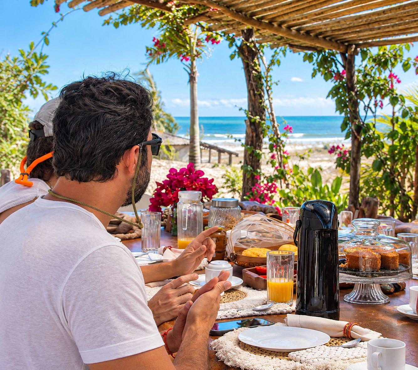 Mesa de comedor al aire libre con vista al océano, con alimentos para el desayuno y una cafetera.