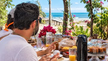 Mesa de comedor al aire libre con vista al océano, con alimentos para el desayuno y una cafetera.