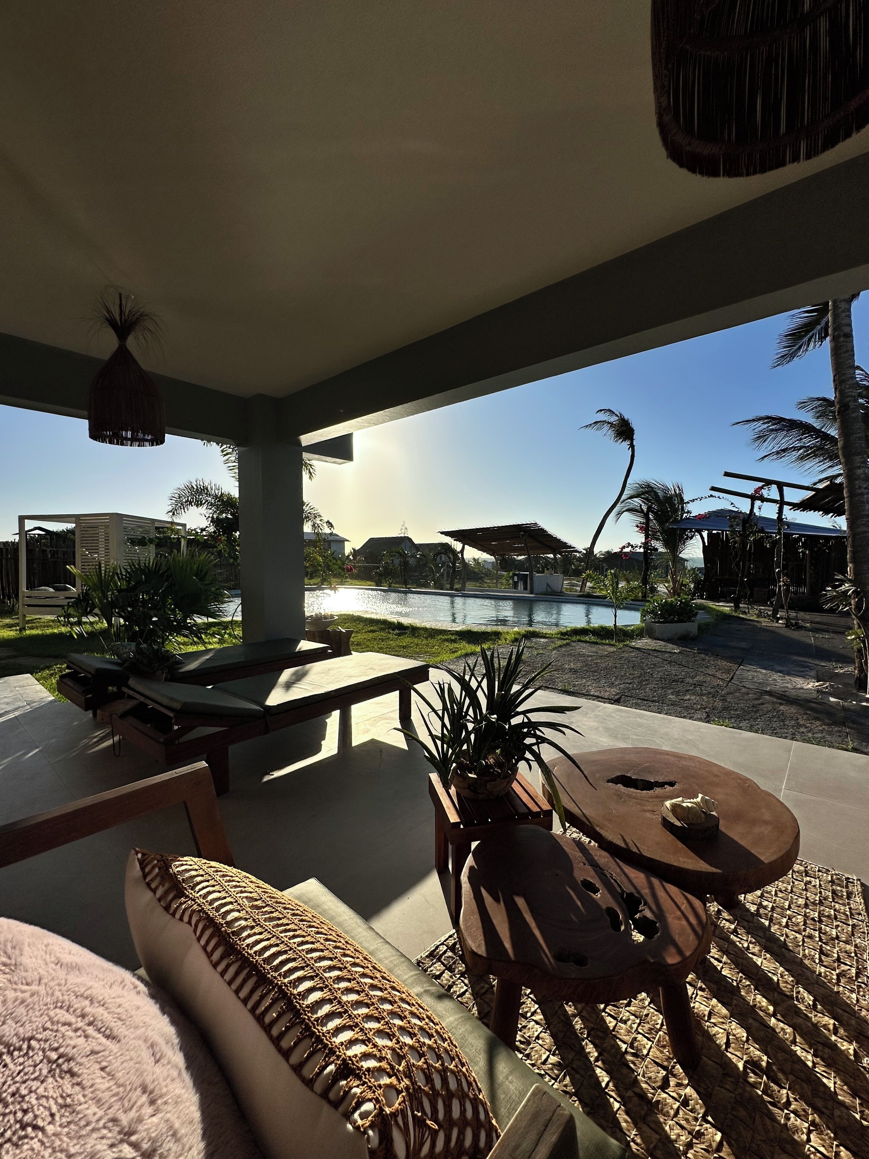 A sunlit outdoor seating area with a pool view, lounge chairs, and a wooden table.