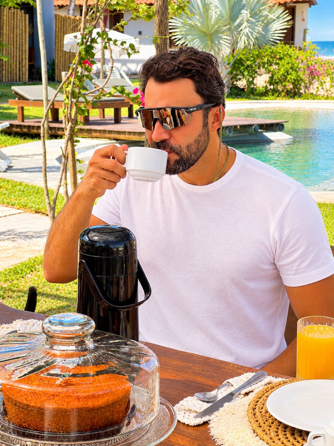 A person seated at a table with a coffee pot, a glass of orange juice, and a covered dessert on a tray.