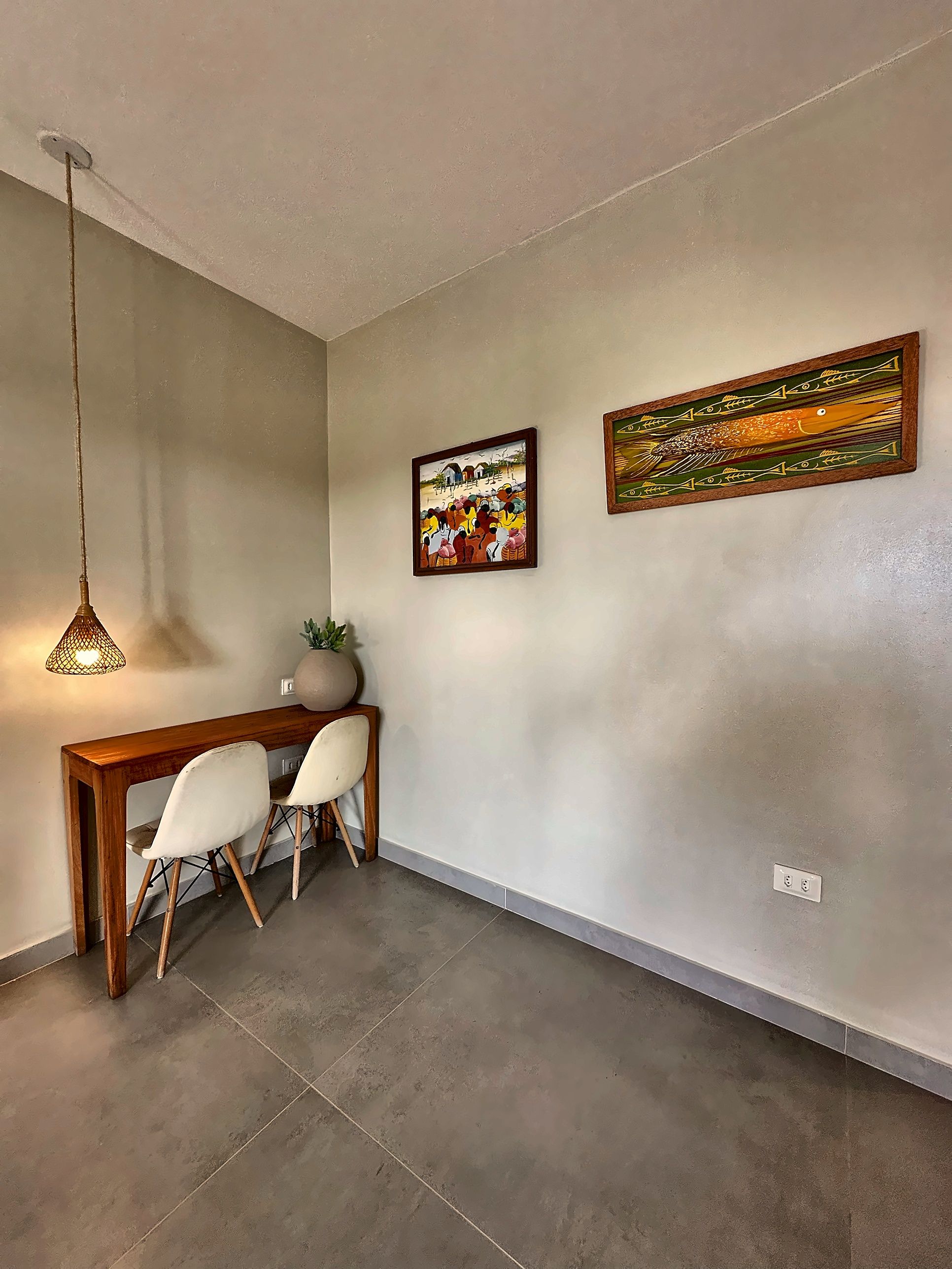 A dining area with a wooden table, two white chairs, a hanging light fixture, and two framed artworks on the wall.