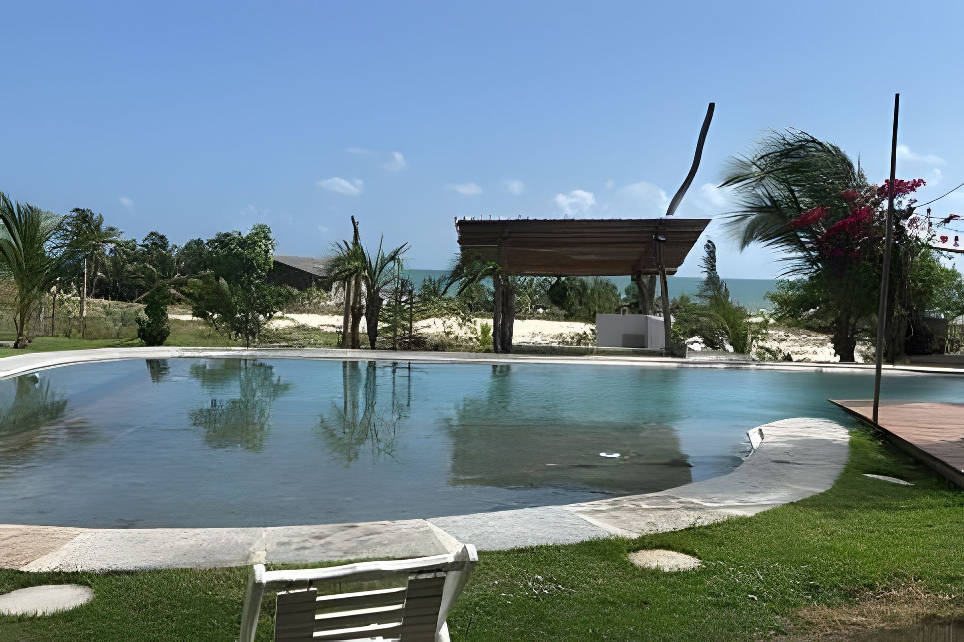 A swimming pool with a wooden deck and a covered seating area, surrounded by greenery and a view of the sea.