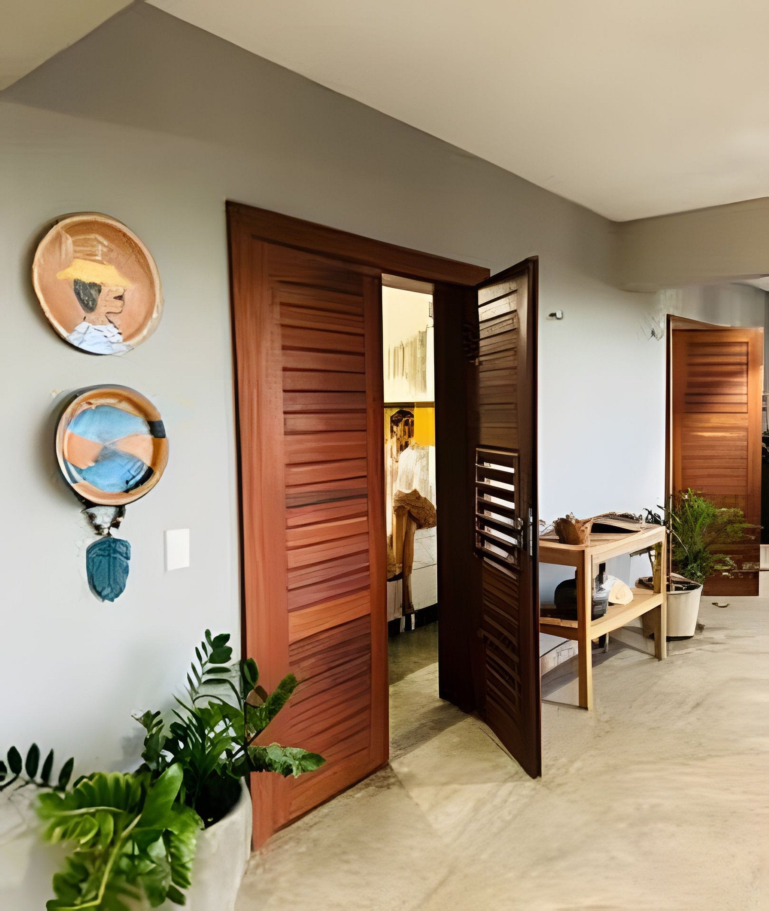 Une porte en bois ouverte donne sur une pièce où se trouvent un lit et un bureau, ainsi qu’une plante en pot et deux objets décoratifs accrochés au mur.