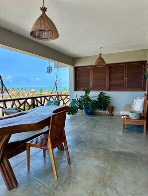 A spacious balcony with a wooden table and chairs, a hanging light fixture, and a view of the ocean. - refugio prea (Cruz)