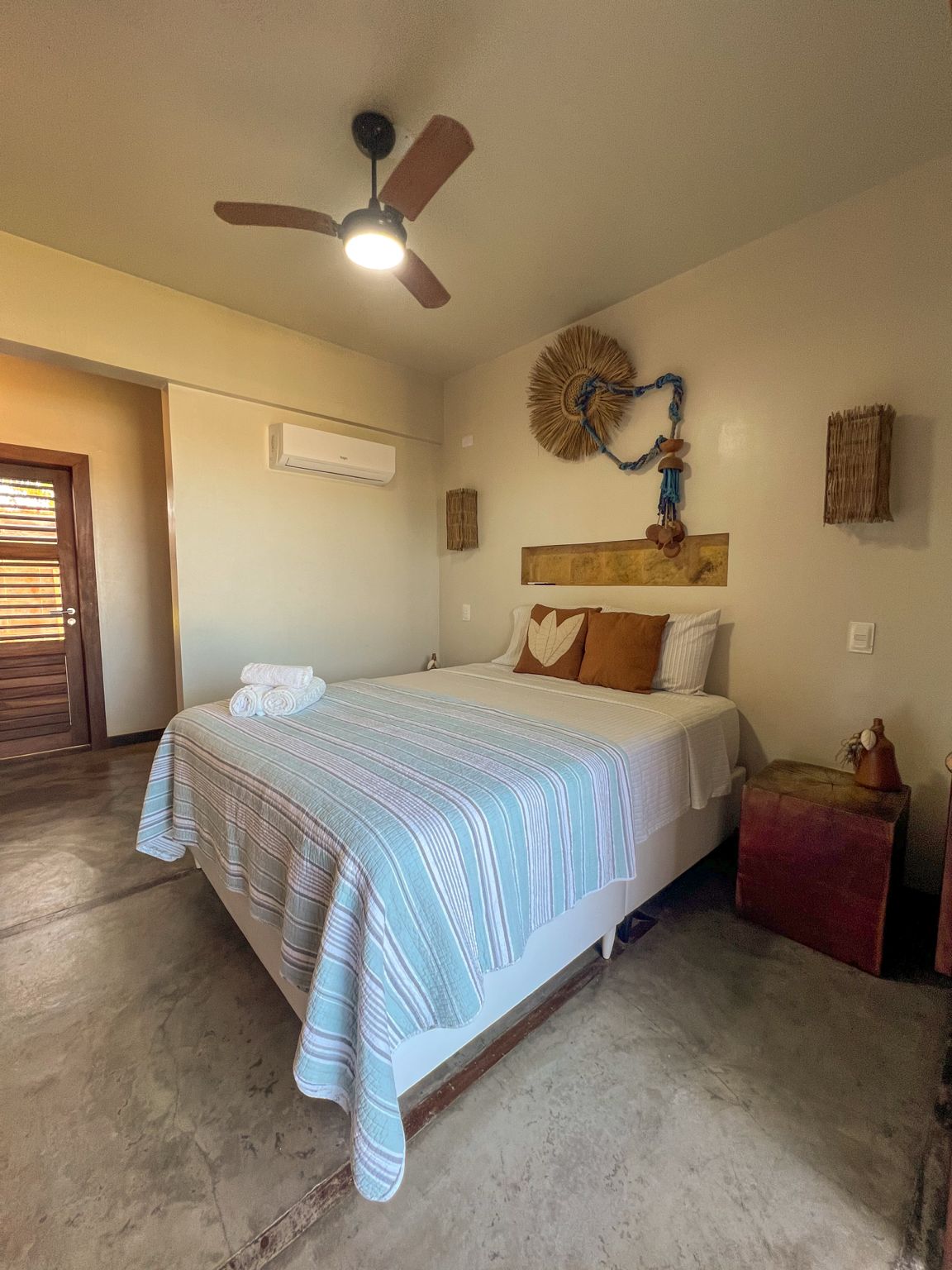 A bedroom with a bed, a ceiling fan, a wall-mounted air conditioner, and a decorative wall hanging.