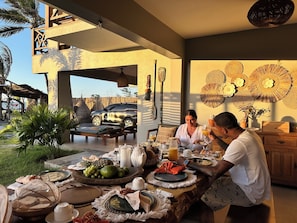 An outdoor dining area with a table set for two, surrounded by tropical plants and a view of a vehicle. - refugio prea (Cruz)