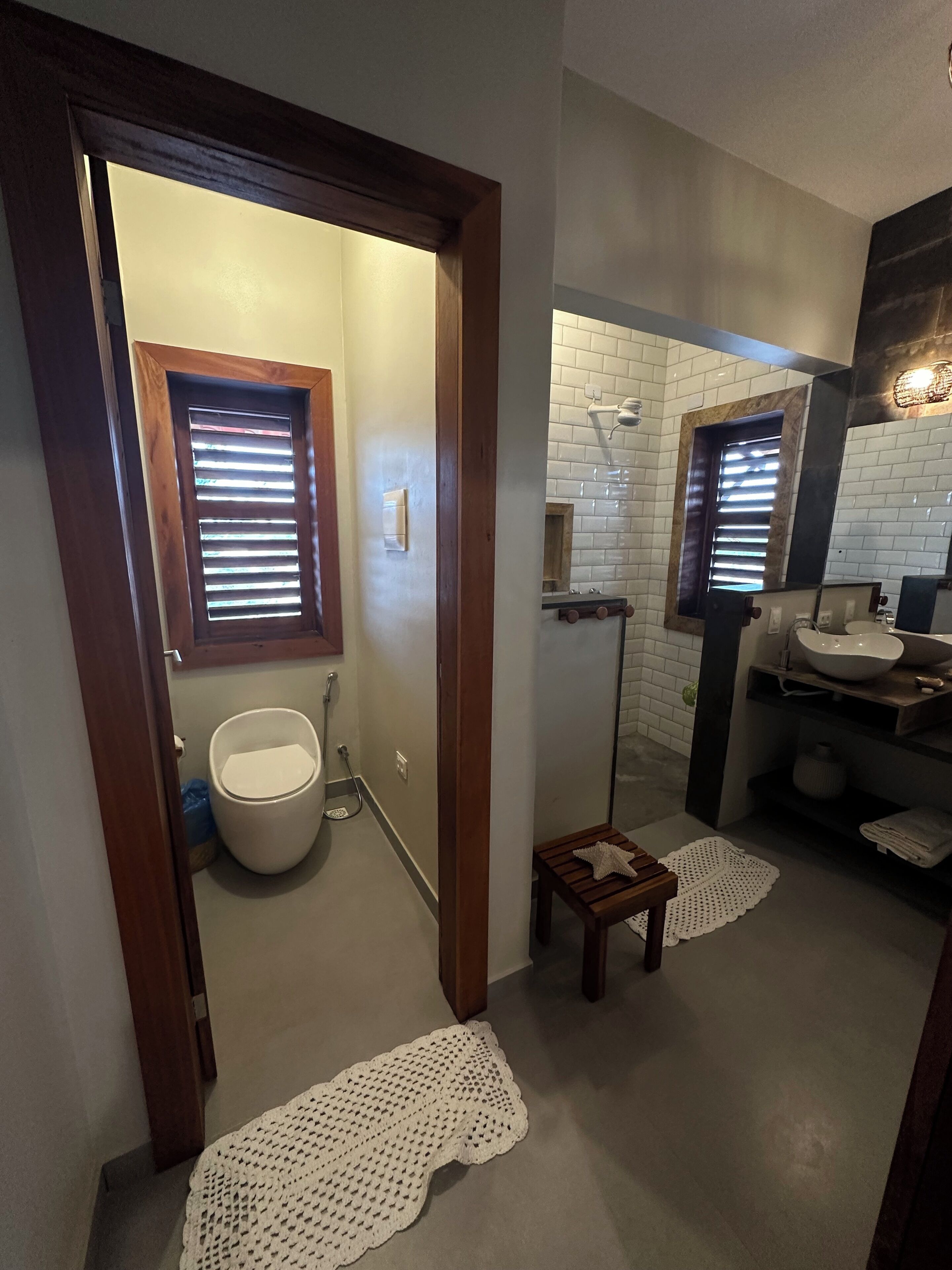 Une salle de bain comprenant des toilettes, un bidet et un lavabo avec un miroir. Il y a deux fenêtres équipées de stores, un porte-serviettes et un petit tabouret en bois.