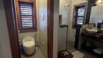 Une salle de bain comprenant des toilettes, un bidet et un lavabo avec un miroir. Il y a deux fenêtres équipées de stores, un porte-serviettes et un petit tabouret en bois.