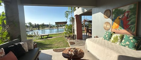 A modern living room with a sofa, a coffee table, and a painting of parrots. There is a view of a swimming pool and a patio area with outdoor furniture.