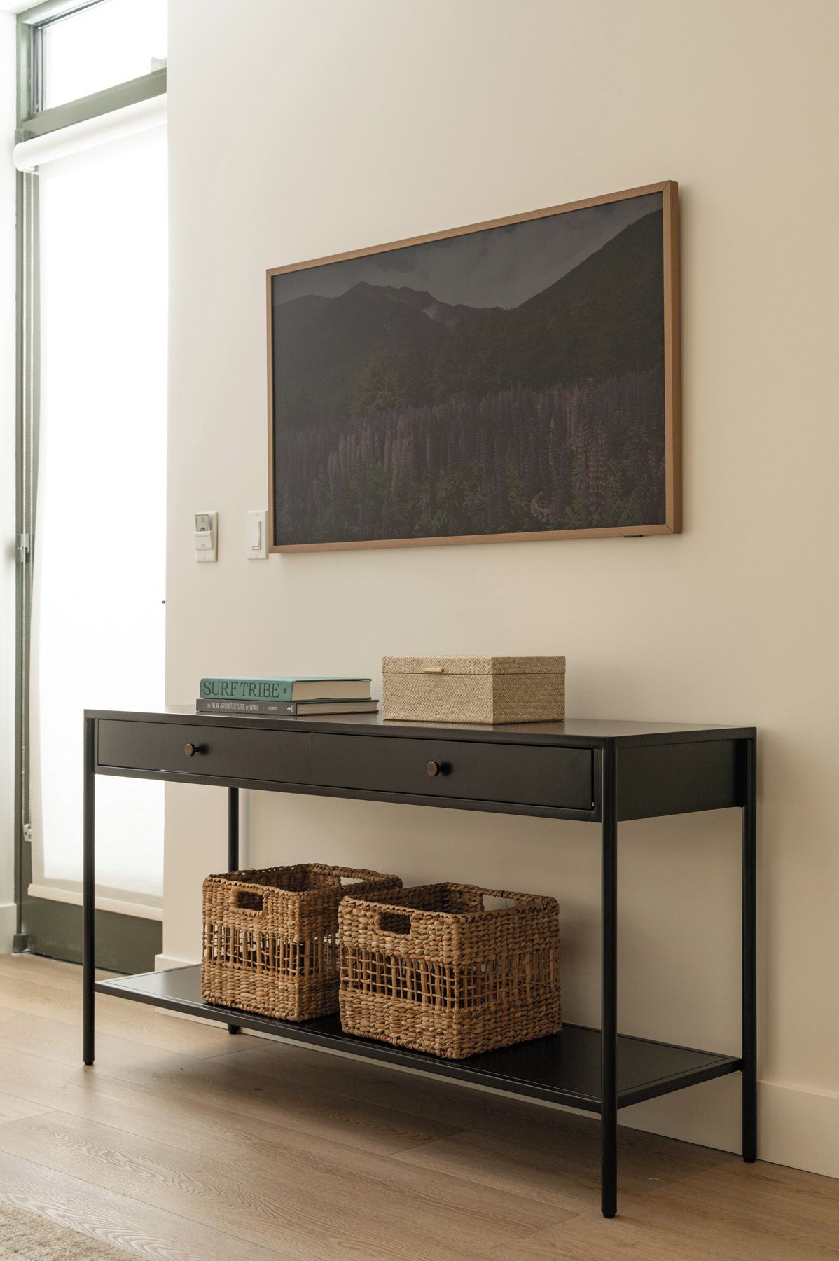 A modern room with a black console table, wicker baskets, a framed landscape painting, and a window with a view.