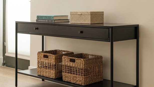 A modern room with a black console table, wicker baskets, a framed landscape painting, and a window with a view.