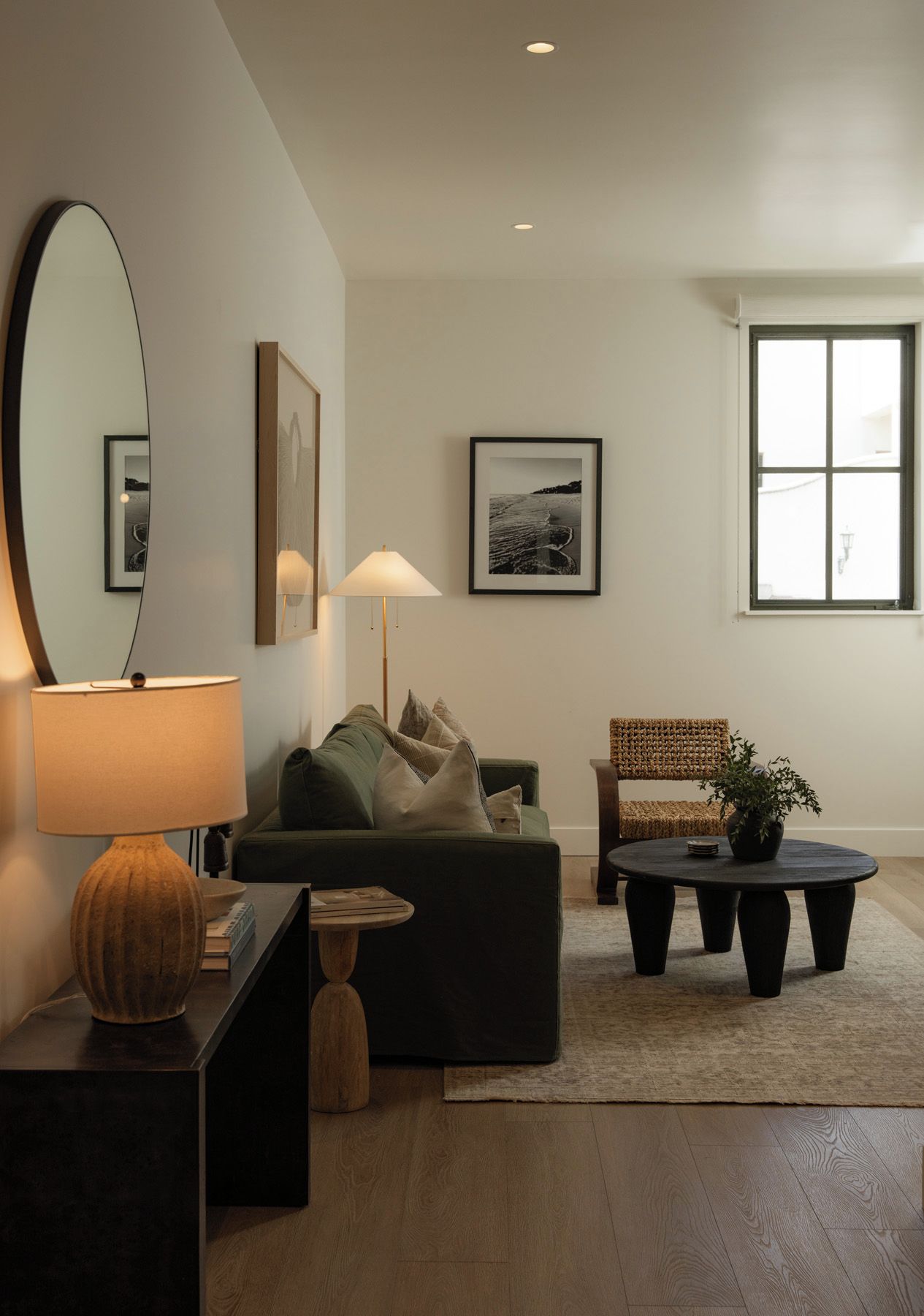A modern living room with a sofa, a round coffee table, a side table with a lamp, and a chair. There is a large mirror on the wall and a framed picture above the sofa.