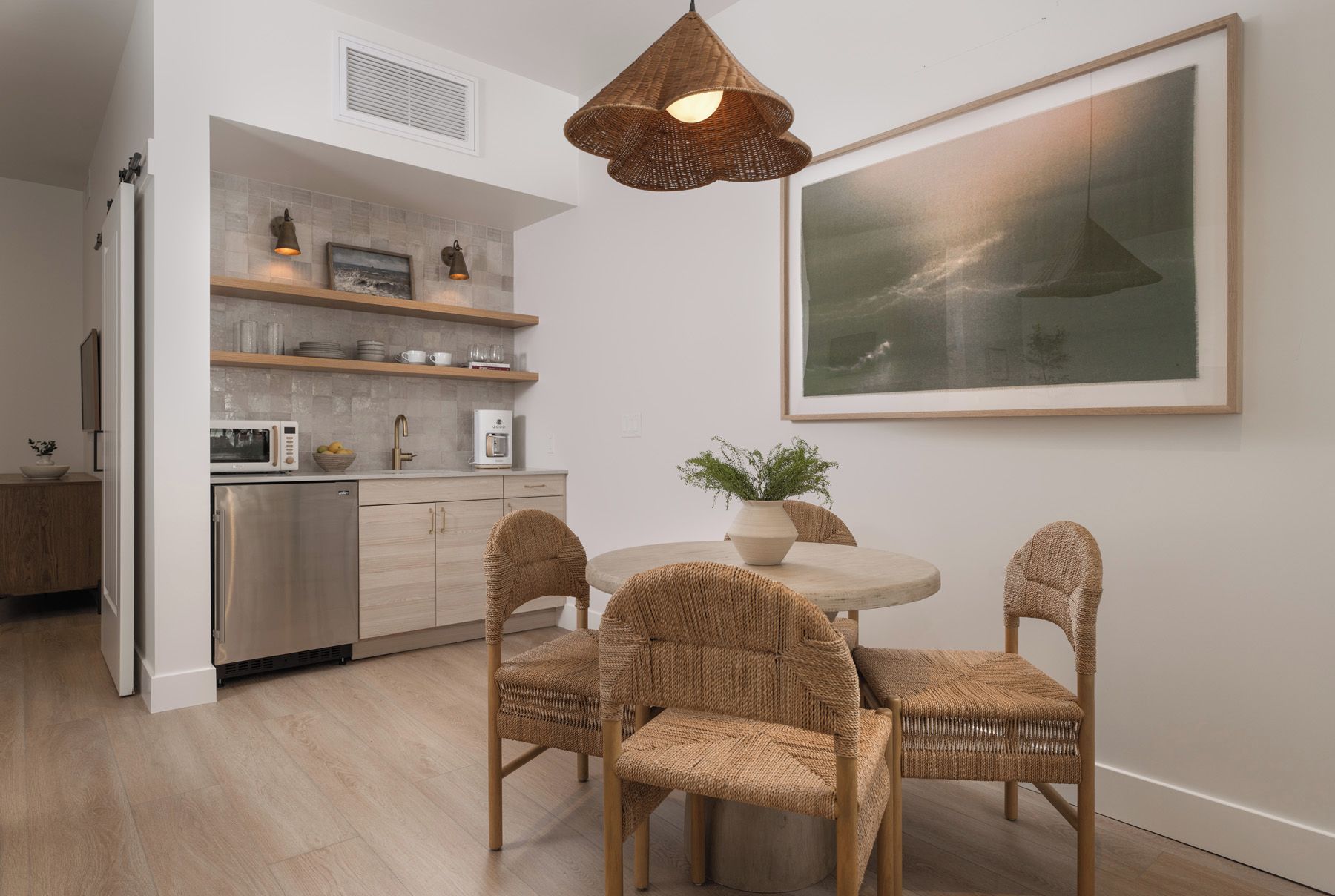 A modern kitchen with a dining area, featuring a round table with four wicker chairs, a hanging light fixture, and a large framed artwork on the wall.