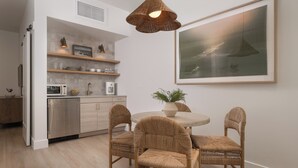 A modern kitchen with a dining area, featuring a round table with four wicker chairs, a hanging light fixture, and a large framed artwork on the wall.