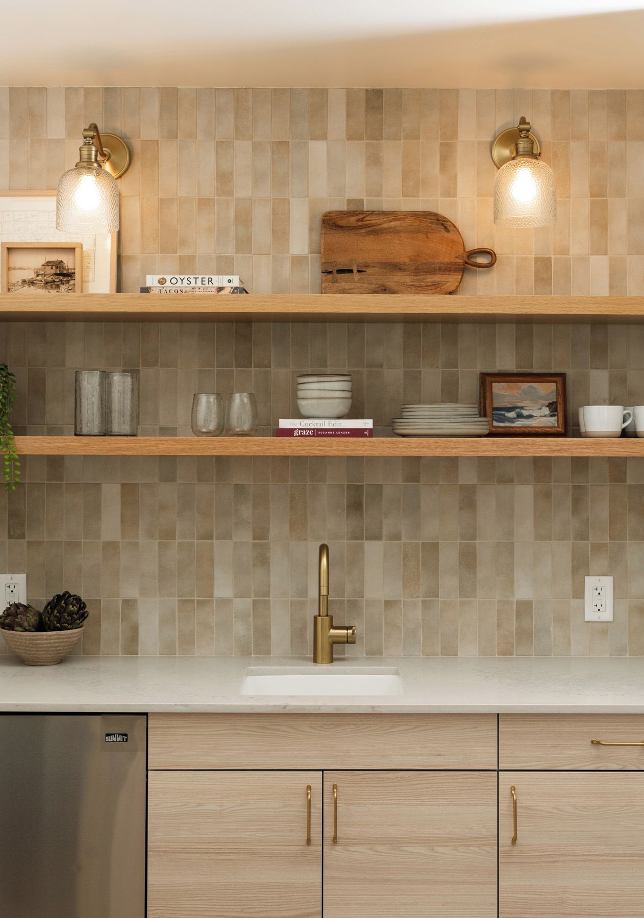 A kitchen with a tiled backsplash, wooden shelves, a stainless steel dishwasher, and a gold faucet.