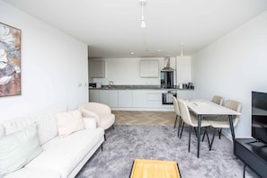 A modern living space with a sofa, dining table, chairs, a TV, and a kitchen area with appliances. - Sleek 2-Bed Salford Apartment near City Centre (Salford)