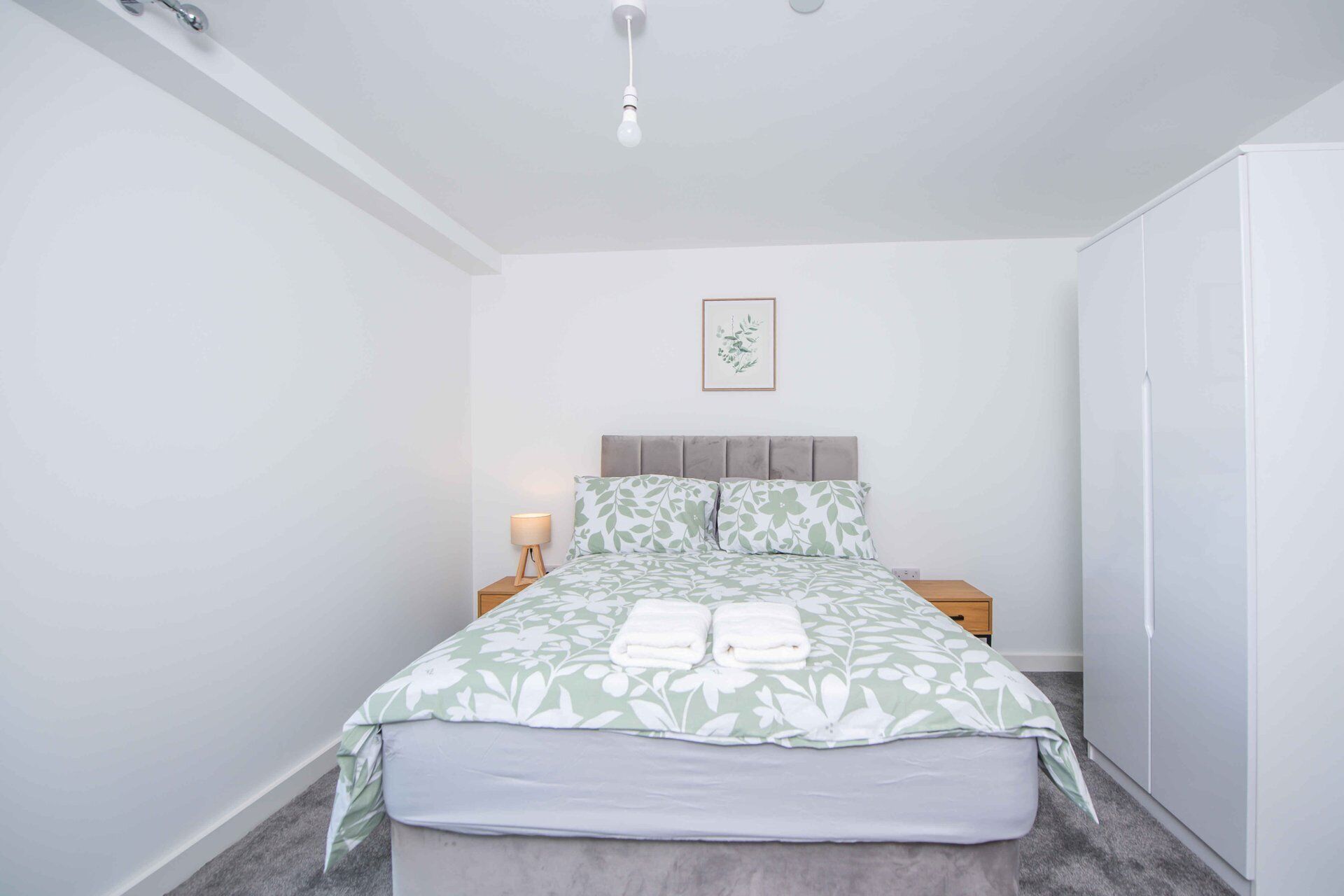 Ein Schlafzimmer mit Bett, Nachttischen, einer Lampe, einem Kleiderschrank und einem gerahmten Bild an der Wand.