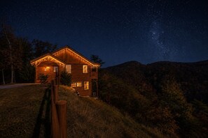 Eine Holzhütte mit Veranda und Lichterkette, umgeben von einer Hügelslandschaft und einem sternenübersäten Nachthimmel.