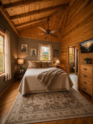 Ein Schlafzimmer in einer Holzhütte mit Bett, Deckenventilator, Fenster mit Vorhängen und einem Bild an der Wand.