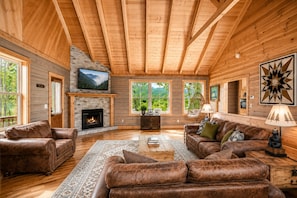 A log cabin interior with a fireplace, leather sofas, a television, and a large window overlooking greenery. - Marvelous Views! Hot tub, Air Hockey, Location, Fire Pit- This Cabin Has It All! (Sevierville)