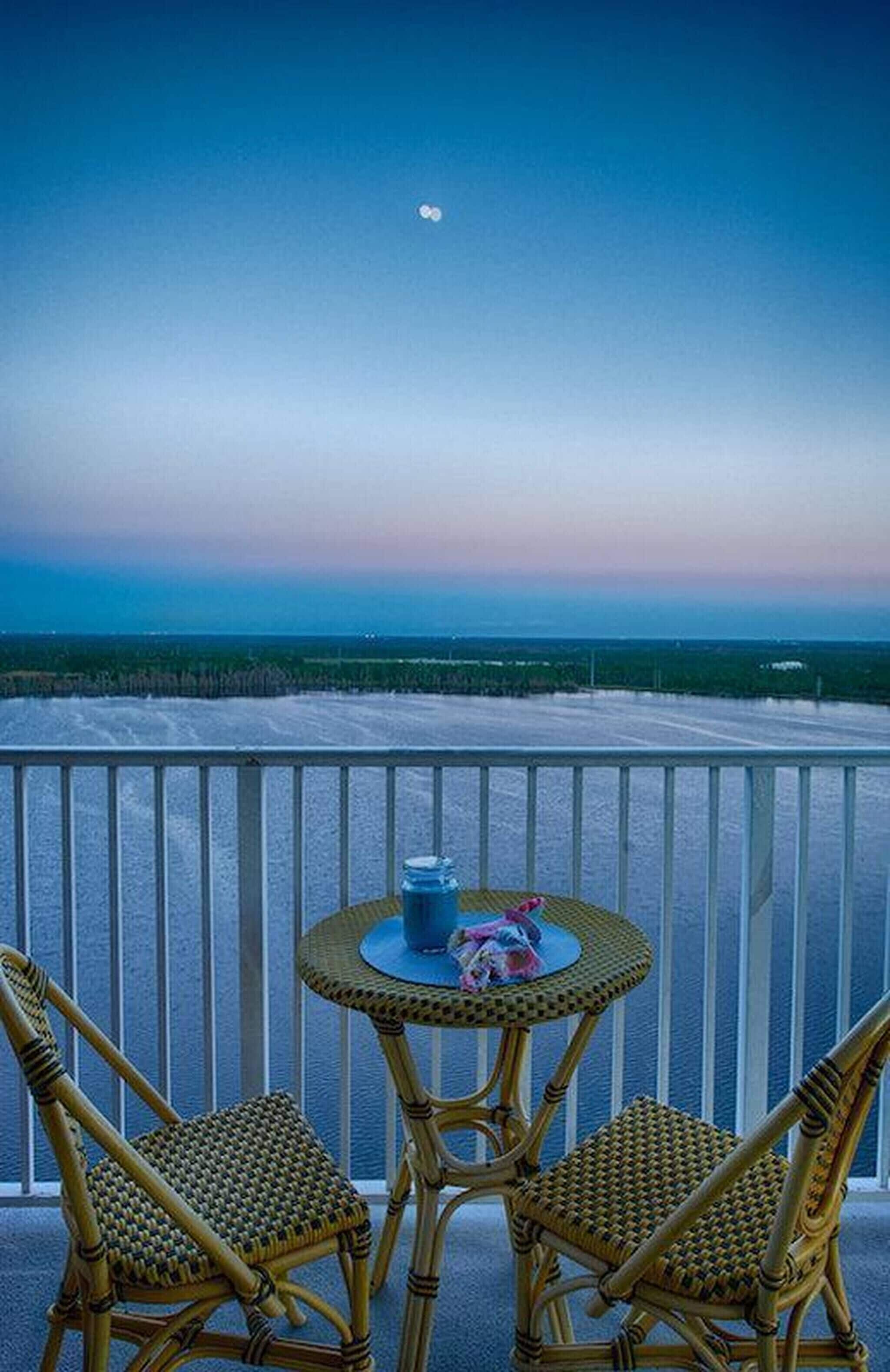 A balcony with two wicker chairs and a small round table, overlooking a beach and ocean.