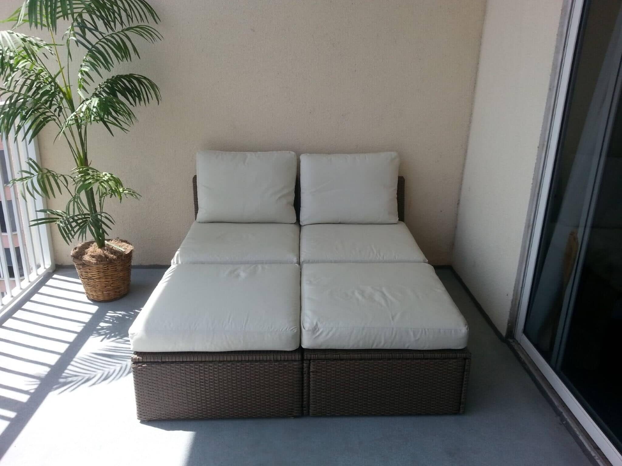 A balcony with a wicker sofa bed, a potted plant, and a view of the outside.
