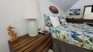 Une chambre à coucher avec un lit, une table de chevet avec une lampe, une sculpture en bois et un mur orné d’un tableau.