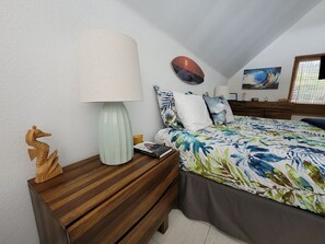 A bedroom with a bed, a nightstand with a lamp, a wooden sculpture, and a wall with a painting. - Hale Kailani - Perfect Oceanfront Home for Relaxing Stays - Sunrise Views (Keaau)