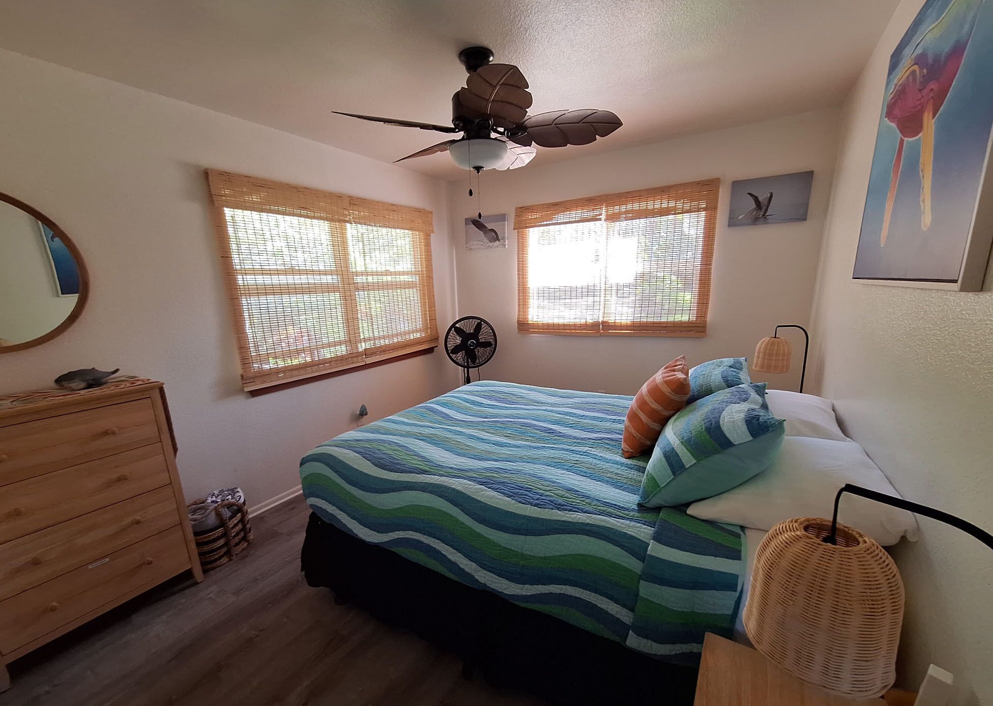 Ein Schlafzimmer mit einem Bett, einem Deckenventilator, zwei Fenstern mit Jalousien, einer Kommode und einem an der Wand befestigten Ventilator.