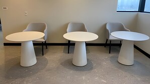 Huoneessa on kolme valkoista pöytää ja niihin sopivat tuolit. Huoneen seinä on vaalea ja siinä on yksi ikkuna.